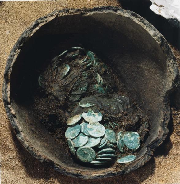 A photograph of an excavated broken ceramic pot containing two clusters of corroded coins with some very degraded organic remains